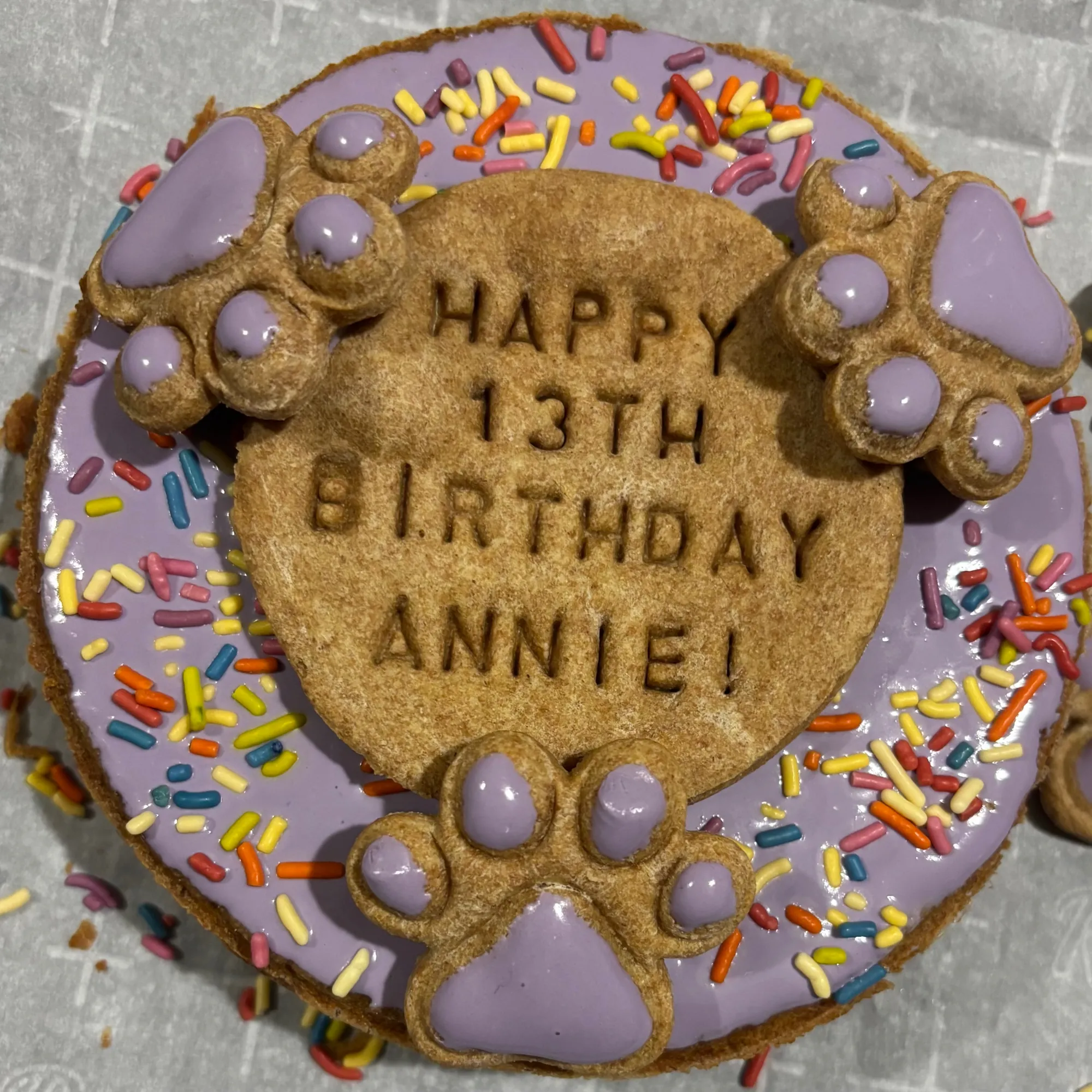 Organic dog birthday cake with purple icing, rainbow sprinkles, and piped paw print decorations